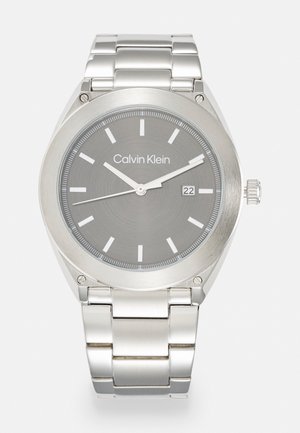Montre en acier inoxydable argenté avec un cadran gris, index horaires blancs, fenêtre de date à 3 heures et un bracelet en métal poli.