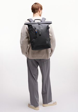 Henkilö, jolla on beige neule ja harmaat housut, kantaa mustaa rullattavaa reppua, jossa on teksti "GOT BAG", selin kameraan.