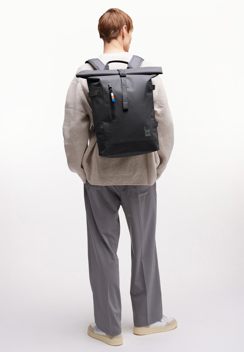 Persona que lleva un suéter beige y unos pantalones grises, cargando una mochila enrollable negra con la etiqueta "GOT BAG", mientras está de espaldas a la cámara.