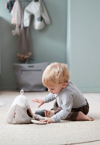 Zacht knuffeldier in een abstracte bergvorm, lichtgrijs en beige met een gat aan één kant. Kind zittend op een geweven tapijt speelt ermee.