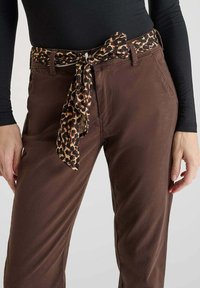 Pantaloni marroni con vestibilità relaxed, dotati di una cintura di tessuto con stampa leopardata legata in vita. Si può notare una texture liscia e una leggera lucentezza.