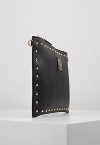 Borsa in pelle nera con borchie dorate lungo i bordi, dotata di chiusura a zip superiore e un logo dorato sul fronte. Design testurizzato ed elegante.