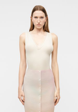 Kobieta o prostych brązowych włosach, ubrana w bezrękawnikowy biały prążkowany top z koronkową lamówką oraz spódnicę z pastelowym gradientem i guzikami, stojąca na tle jednolitego tła.