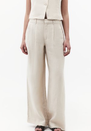 Frau trägt beige Leinenhose mit hohem Bund und weitem Bein, kombiniert mit einer passenden kurzen Weste und schwarzen Sandalen mit offenen Zehen.