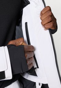 Valge veekindel jakk mustade detailidega, millel on tõmblukuga sisetasku ja tekstureeritud must randmeosa. Käed haaravad kangast.