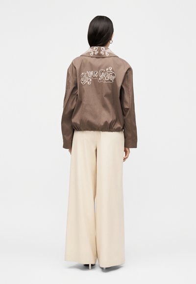 Γυναίκα με πλάτη, φορά καφέ ζακέτα με λευκή κέντημα στην πλάτη και μπεζ παντελόνι με φαρδιά πόδια.