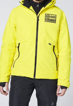 Mand iført en skarp gul jakke med lynlås og sort logo "PLUSMINUS" på brystet samt sorte lynlåslommer, kombineret med sorte bukser.