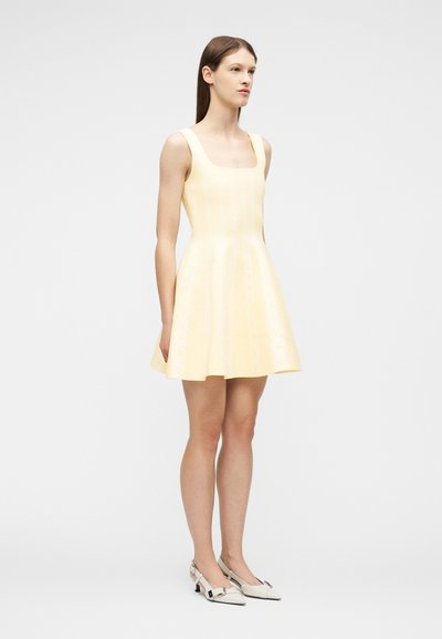 Νέα γυναίκα που φορά αμάνικο ανοιχτό κίτρινο φόρεμα με φουσκωτή φούστα και λευκά παπούτσια με χαμηλό τακούνι, στέκεται μπροστά σε ένα απλό ανοιχτό φόντο.