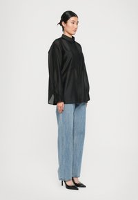 Kvinne som står i profil iført en løs svart satengskjorte, lyseblå rette jeans og svarte spisse høye hæler.
