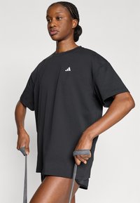 Μαύρο υπερμεγέθες μπλουζάκι από απαλό ύφασμα, με μικρό λευκό λογότυπο στο στήθος. Το μοντέλο κρατά γκρι λαστιχένιες ταινίες αντίστασης σε κάθε χέρι.