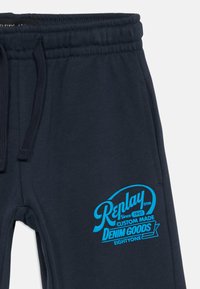 Pantalones de chándal azul marino con cintura elástica y cordón, que presentan un logo en brillante azul de "Replay Custom Made Denim Goods" en el muslo.