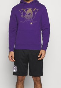Sudadera morada con un diseño gráfico dorado en el frente, tela de mezcla de algodón y bolsillo canguro. Combinada con pantalones cortos de malla negros.