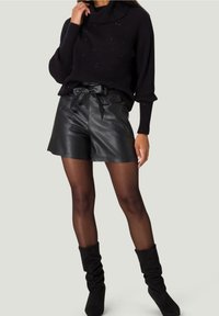 Mujer con un suéter de cuello alto de canalé negro, pantalones cortos de cuero negro de cintura alta con cinturón, medias transparentes negras y botas negras holgadas.