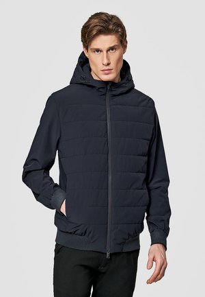 Młody mężczyzna ubrany w czarną pikowaną kurtkę zapinaną na zamek z kapturem i ściągaczami na mankietach, stojący na tle jednolitego jasnoszarego tła.