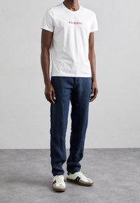 Hvid bomulds t-shirt med "PIZZICATO" i rød, parret med mørkeblå linnedbukser og hvide sneakers med sorte striber og grønne accenter.