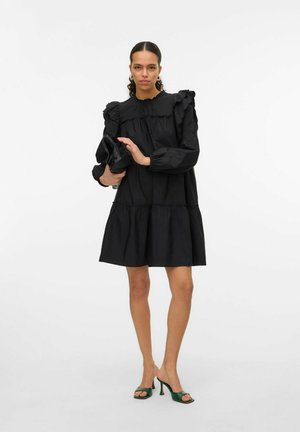 Black dress with ruffled shoulder details, long sleeves, tiered design, and a relaxed silhouette. Paired with green heeled sandals.