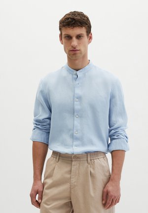 Homme portant une chemise bleu clair à col mandarin avec manches retroussées et pantalon beige, debout devant un fond blanc uni.