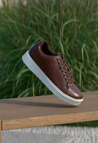 Baskets en cuir marron avec une texture lisse, semelle en caoutchouc blanche et bout rond. Présente un laçage à l'avant et un design minimaliste.