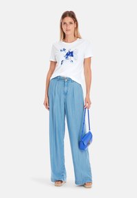 White t-shirt with blue fish graphic, paired with light blue wide-leg jeans. Model also carries a small blue handbag. Natural fabric texture.