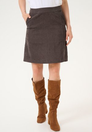 Femme portant une jupe marron mi-longue avec des poches et des bottes à talons en suédine marron, se tenant sur un fond blanc.