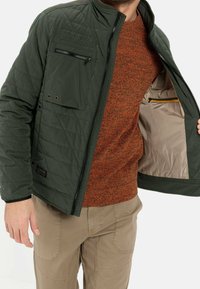 Quiltet grøn jakke med lynlåslukket brystlomme og beige for, båret over en struktureret orange sweater og brune bukser. Har en opretstående krave.