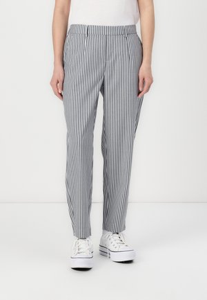 Person iført grå og hvide bukser med lodrette striber, forlommer og hvide plateau-sneakers, stående mod en hvid baggrund.