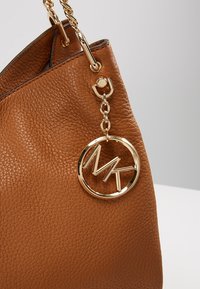 Sac en cuir marron avec une finition texturée, doté d'une chaîne en or et d'un charme circulaire portant les initiales "MK".