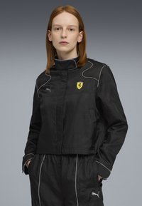 Jachetă neagră scurtă cu guler înalt, având cusături albe și un logo Ferrari. Materialul pare neted și texturat.