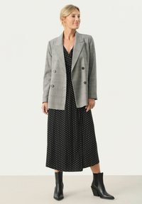 Grau karierte, doppelt knöpfbare Blazer über einem schwarzen Polka-Dot-Kleid, knöchellang, mit schwarzen, hohen Stiefeletten. Glatte Texturen und maßgeschneiderte Passform.