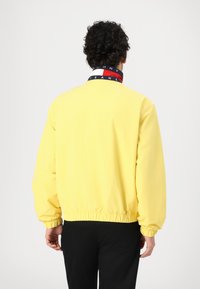 Gelber Pulloverjacke mit elastischen Bündchen und Saum, ausgestattet mit einem mehrfarbigen Kragen mit Text. Glattes Material, entspannte Passform.