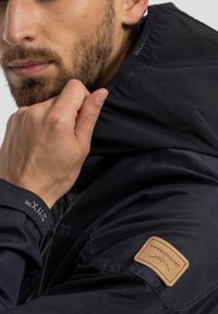 Giacca nera impermeabile con colletto alto, tessuto tattile e una toppa in pelle marrone sulla spalla con l'etichetta "Swedemount."