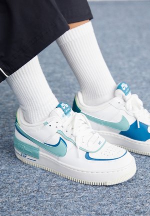 Personne portant des baskets Nike Air Force 1 blanches avec des accents bleu clair et sarcelle et des chaussettes blanches côtelées sur un tapis bleu.