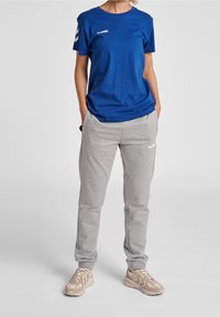 Camiseta de algodón azul con logo blanco y pantalones de chándal grises. Los pantalones tienen una cinturilla elástica y tobillos ajustados, llevados con zapatillas beige.