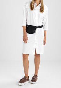 Valkoinen pitkähihaisen painonappitakki, polvenpituinen, edessä halkio. Musta vyölaukku kiinnitettynä, yhdistetty ruskeisiin liukusjalkaimiin.