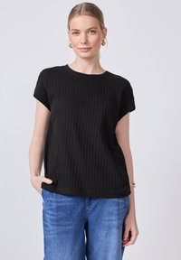 Žena s blond vlasy, která má na sobě zlaté kruhové náušnice, černý pletený top s krátkým rukávem, modré džíny a zlatý náramek, stojí s jednou rukou v kapse.