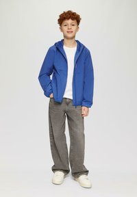 Blau leichte Jacke mit Kapuze, zwei Fronttaschen, getragen über einem weißen Hemd und kombiniert mit grauen Jeans und weißen Sneakers.