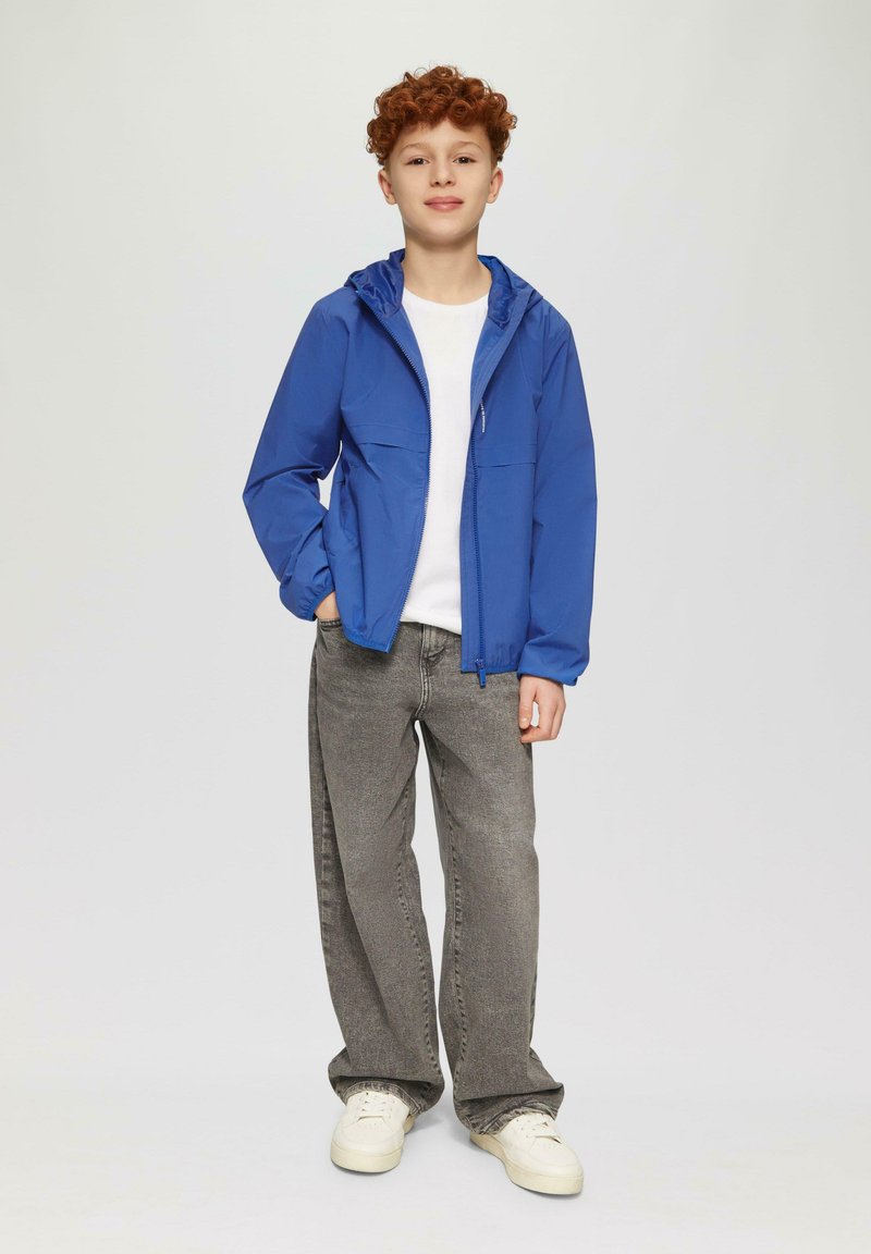 Blau leichte Jacke mit Kapuze, zwei Fronttaschen, getragen über einem weißen Hemd und kombiniert mit grauen Jeans und weißen Sneakers.