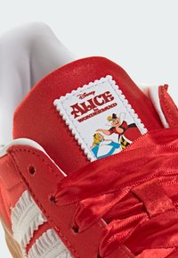 Κόκκινο αθλητικό παπούτσι με λαμπερό σατέν επάνω μέρος, διαθέτει λευκή λαστιχένια σόλα και ετικέτα με χαρακτήρες από το «Η Αλίκη στη Χώρα των Θαυμάτων».