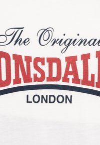 Fehér pamut póló, merész piros és sötétkék szöveges dizájnal. A szöveg így olvasható: "Az Eredeti Lonsdale London", ívelt elrendezésben.