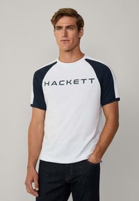 Biely tričko s krátkym rukávom a námorníckymi raglánovými rukávmi, so slovom "HACKETT" vytlačeným tmavomodrou farbou na hrudníku. Voľný strih.