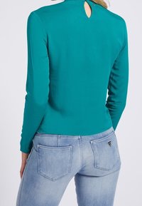 Maglia a maniche lunghe color teal con apertura a chiave sulla schiena; abbinata a jeans denim azzurri con due tasche posteriori e un piccolo logo sulla tasca destra.