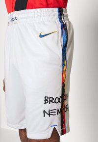 Shorts de basket en blanc de Nike avec tissu en maille, ceinture élastique, rayures latérales bleues et jaunes, et "BROOKLYN NEW YORK" brodé en noir.