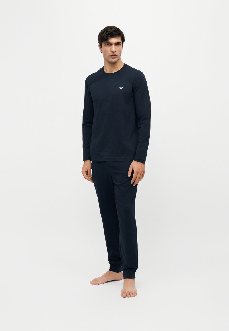 Homme debout pieds nus portant un haut à manches longues bleu marine et un pantalon assorti sur un fond blanc uni.