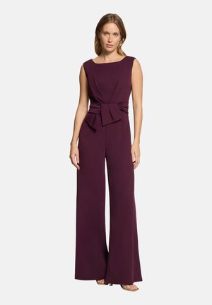 Woman wearing sleeveless dark purple jumpsuit with wide legs and a large fabric belt tied at the waist, standing against a plain background.