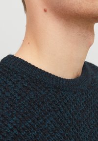 Mörkblå stickad tröja med ett texturerat mönster, rund halsringning och ribbade ärmslut. Tyget verkar mjukt och varmt.