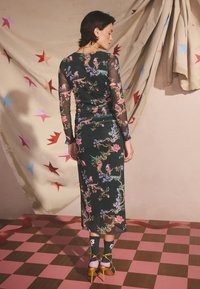 Vestito lungo verde con stampa floreale, maniche nere trasparenti, design aderente e spacco sull'orlo. Indossato con tacchi gialli e calzini a fantasia.