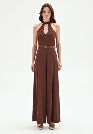 Femme debout portant une combinaison sans manches marron à col licou avec des jambes larges et une ceinture assortie, associée à des talons ouverts dorés.