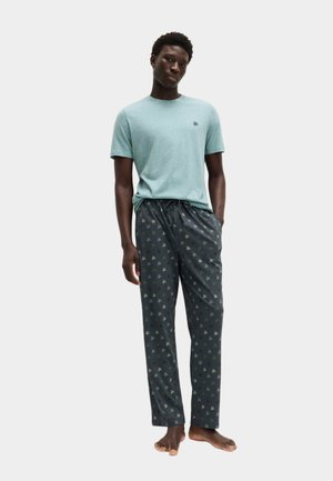 Mężczyzna ubrany w jasnoniebieską koszulę z krótkim rękawem i ciemne spodnie od piżamy w nadruk z motywem palm, stojący boso na jednolitym tle.