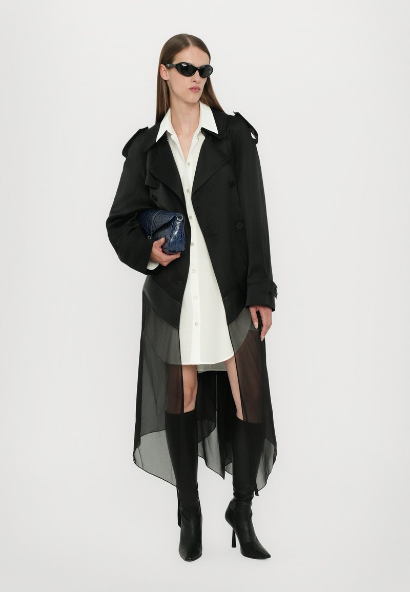 Cappotto trench nero con orlo trasparente, indossato sopra un abito camicia bianco. La modella lo ha abbinato a stivali alti neri con tacco a spillo e occhiali da sole scuri.