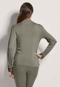 Olivgrünes Langarmoberteil mit hohem Kragen, gerafften Seiten und verzierten Bündchen mit Pailetten. Weicher, elastischer Stoff mit glatter Textur.
